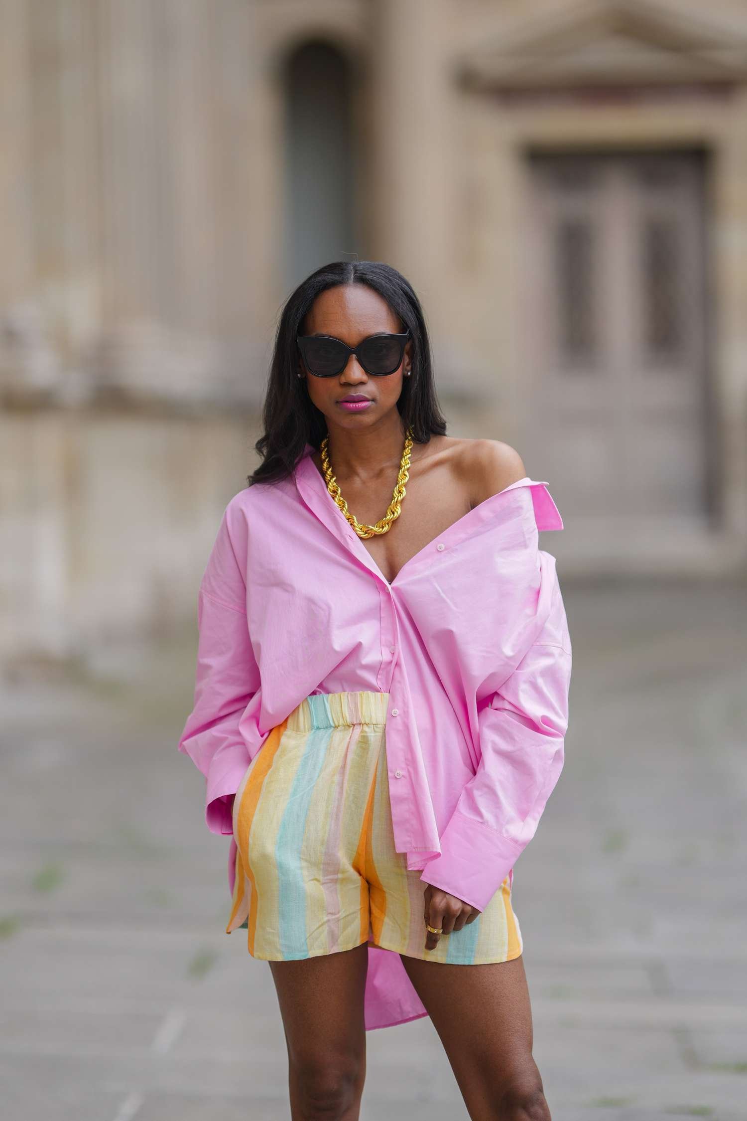 Woman eating away an oversized pink button down, gold chain necklace, and multicolor stripe shorts