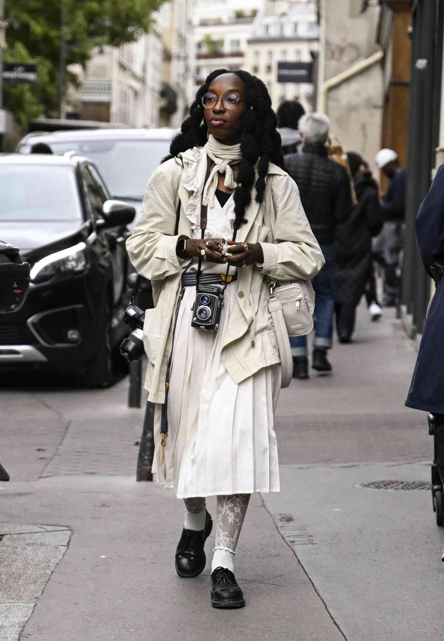 A guest be seen wearing a cream jacket, white dress and black place with white scarf outside the Sacai show during Womenswear Spring/Summer 2025 as component of Paris fashion Week