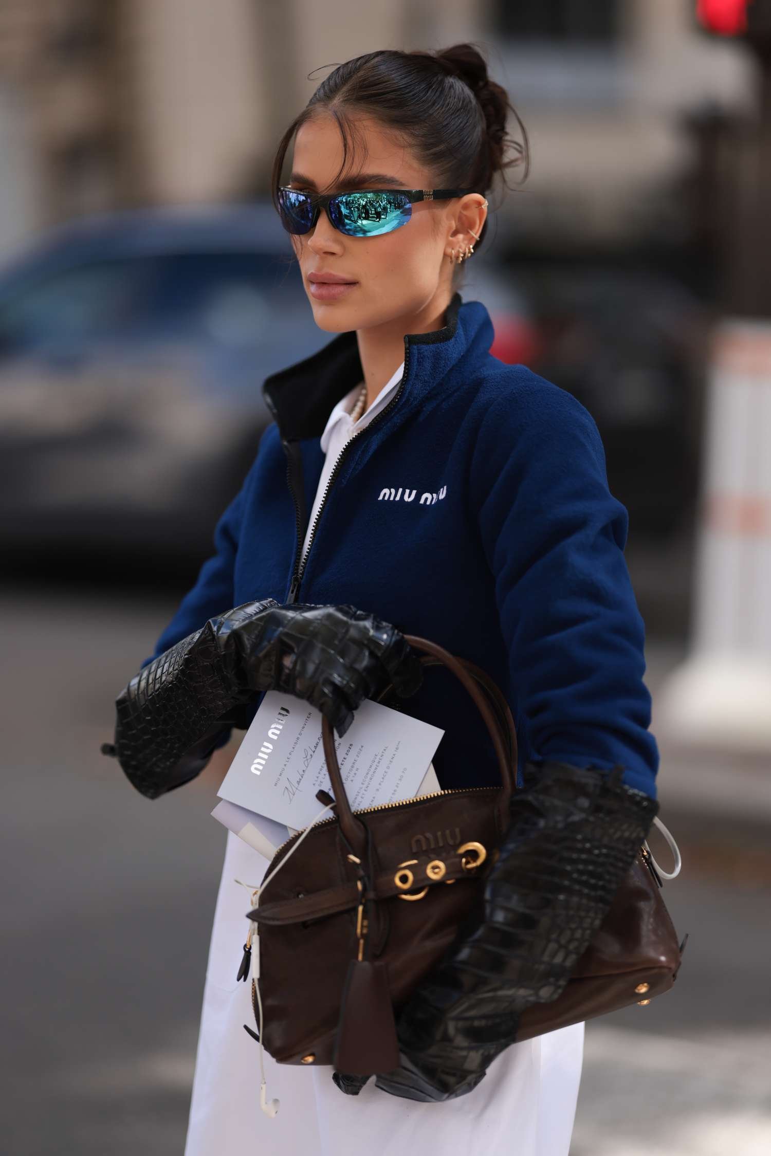 Masha Lobanova is seen wearing several small gold hoop earrings, sportsmanlike black sunglasses with blue mirrored lens and a gold Miu Miu logotype at the temples, A pearl necklace, a cropped dark blue fleece slipover with a black zipper from Miu Miu, pair with a long white shirt underneath also from Miu Miu, long black leather gloves with crocodile embossing, and a rectangular brown leather bag with gold details from Miu Miu before the Miu Miu Womenswear Spring/Summer 2025 Show